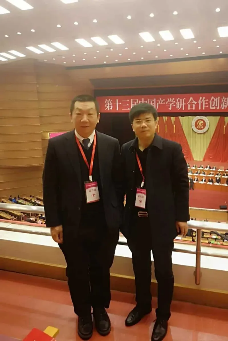 Two adults in dark clothing and red lanyards stand before a conference stage with audience seating behind them.