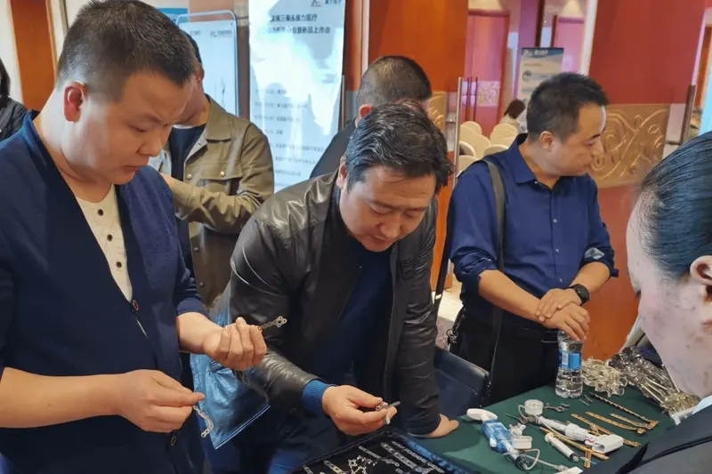 Six main participants examine small metal components and instruments at a display table in a hotel-like meeting room, with additional indistinct figures in the background.