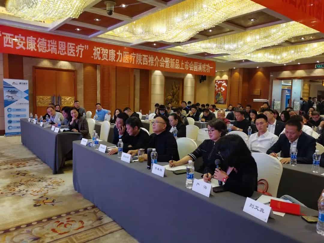 Approximately 35 to 40 attendees sit at long covered tables in a hotel-style function room with name placards, bottled water, and an overhead event banner.