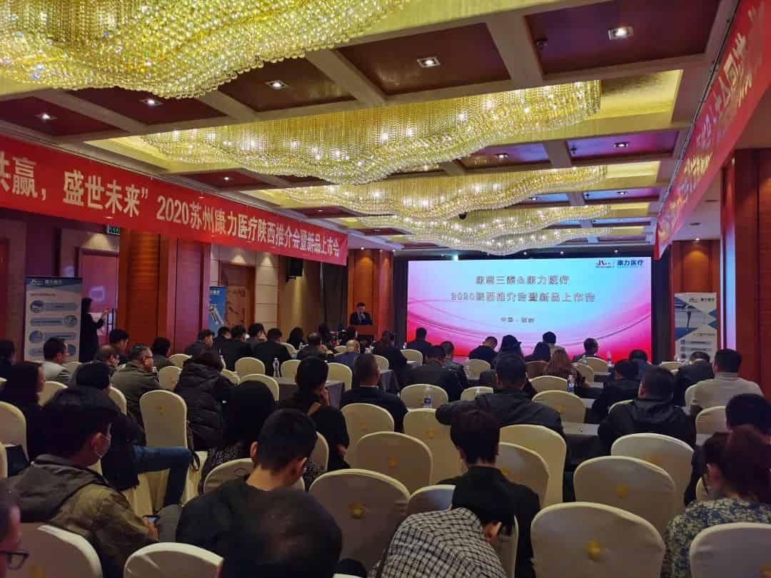Approximately 60 to 70 attendees sit in cream-colored chairs facing a projection screen and a speaker at the front of a conference hall.