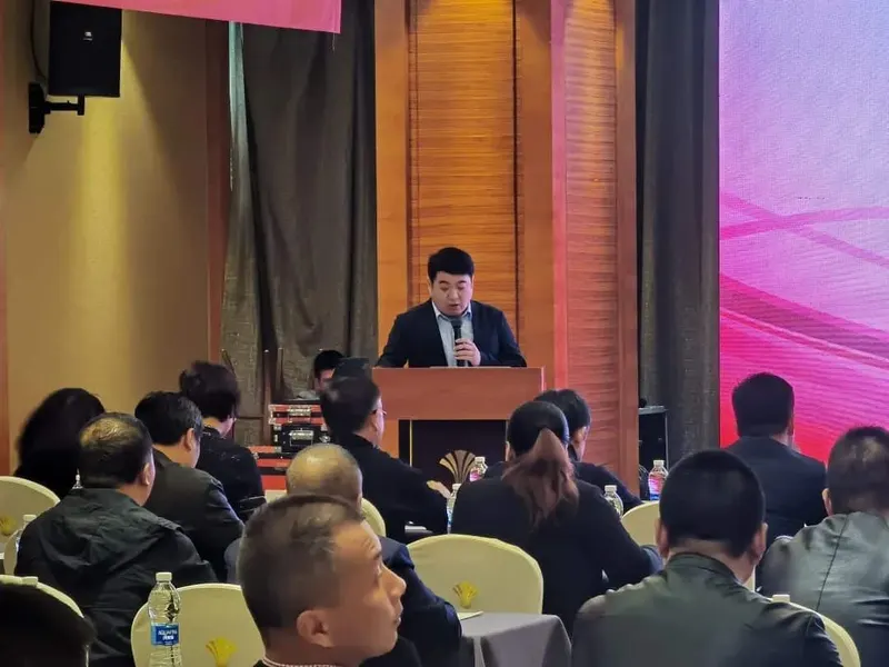 A speaker holds a handheld microphone behind a lectern while approximately fifteen attendees face the front of a decorated conference room.