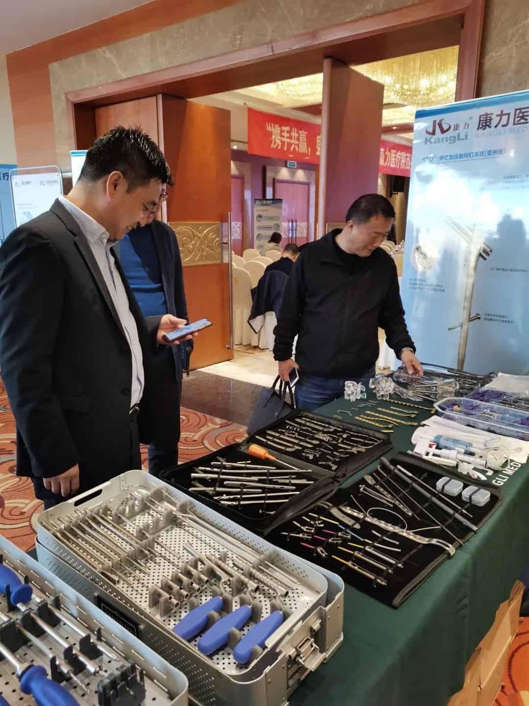 Three participants stand around a table of instruments beside a freestanding banner and event signage in an indoor conference or exhibition venue.