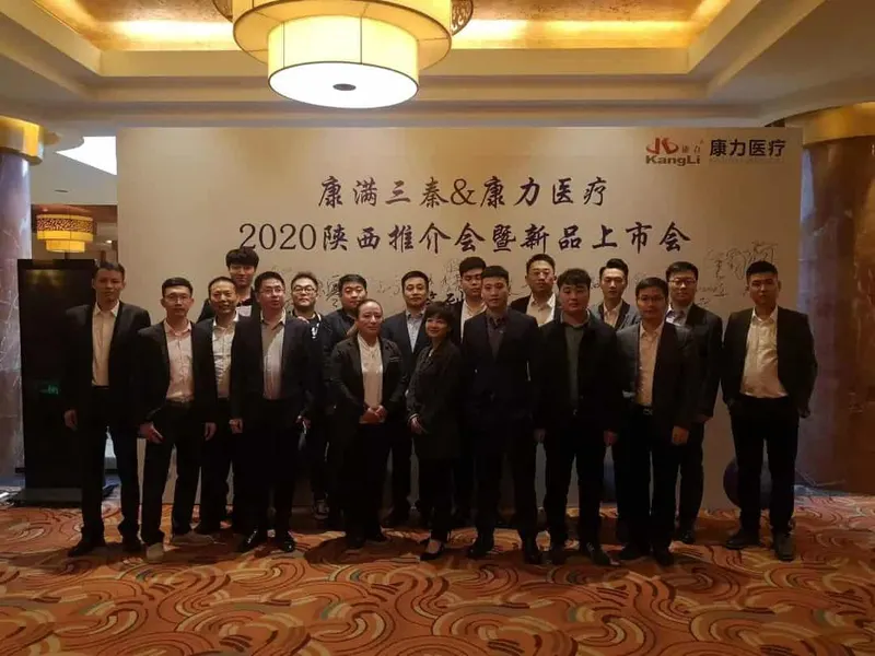 Nineteen participants stand in rows facing the camera before a large cream-colored printed backdrop inside an event venue.