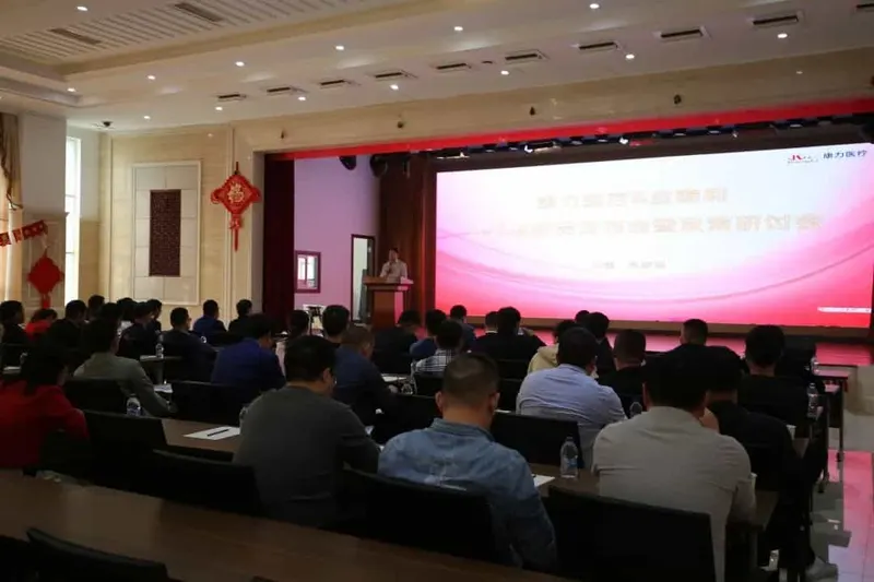 A speaker stands at a lectern on a stage before about thirty seated participants, with a large presentation screen and red stage backdrop.