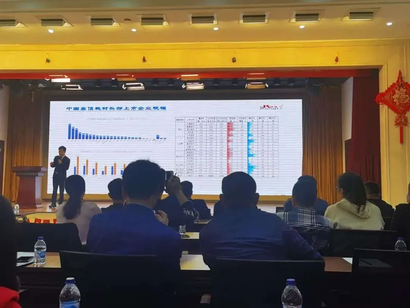 One speaker stands beside a projected presentation screen while about twelve participants sit at tables in a conference room.