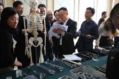 About twelve people gather around tables covered with instruments, papers and trays beside a standing anatomical skeleton in an indoor meeting space.
