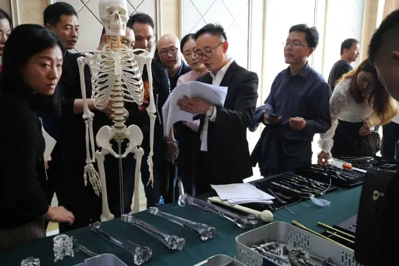 About twelve people gather around tables covered with instruments, papers and trays beside a standing anatomical skeleton in an indoor meeting space.