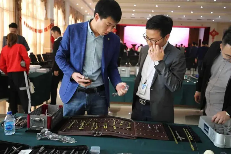 Three people stand at a product table in a conference hall while additional participants examine displayed items near rows of chairs and a presentation screen.