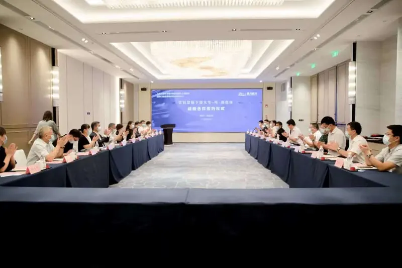 Approximately 25 participants sit around U-shaped draped tables with pink name cards, papers and water bottles beneath a blue presentation screen.