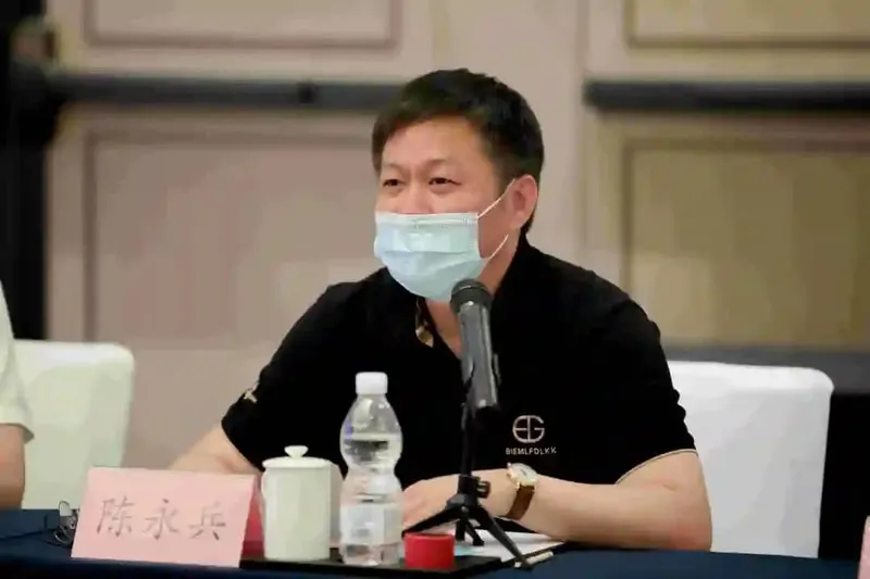 A seated participant in a black polo shirt and pale blue surgical mask faces a tabletop microphone at a conference table.