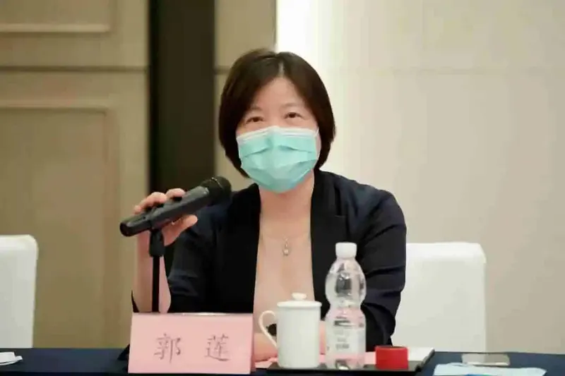 A seated participant in a black jacket and light teal surgical mask holds a handheld microphone beside a name card and bottled water.