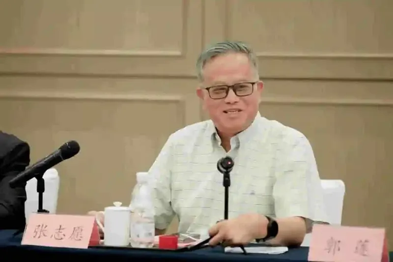 A seated participant wearing glasses and a patterned shirt faces a tabletop microphone at a conference table with documents and refreshments.