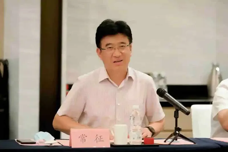 A seated participant in a pale pink collared shirt and glasses faces a tabletop microphone, with a blank projection screen behind.