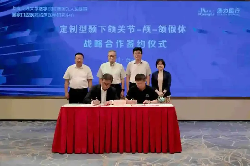 Two seated participants sign separate papers with black pens while four participants stand behind them beneath a presentation screen.