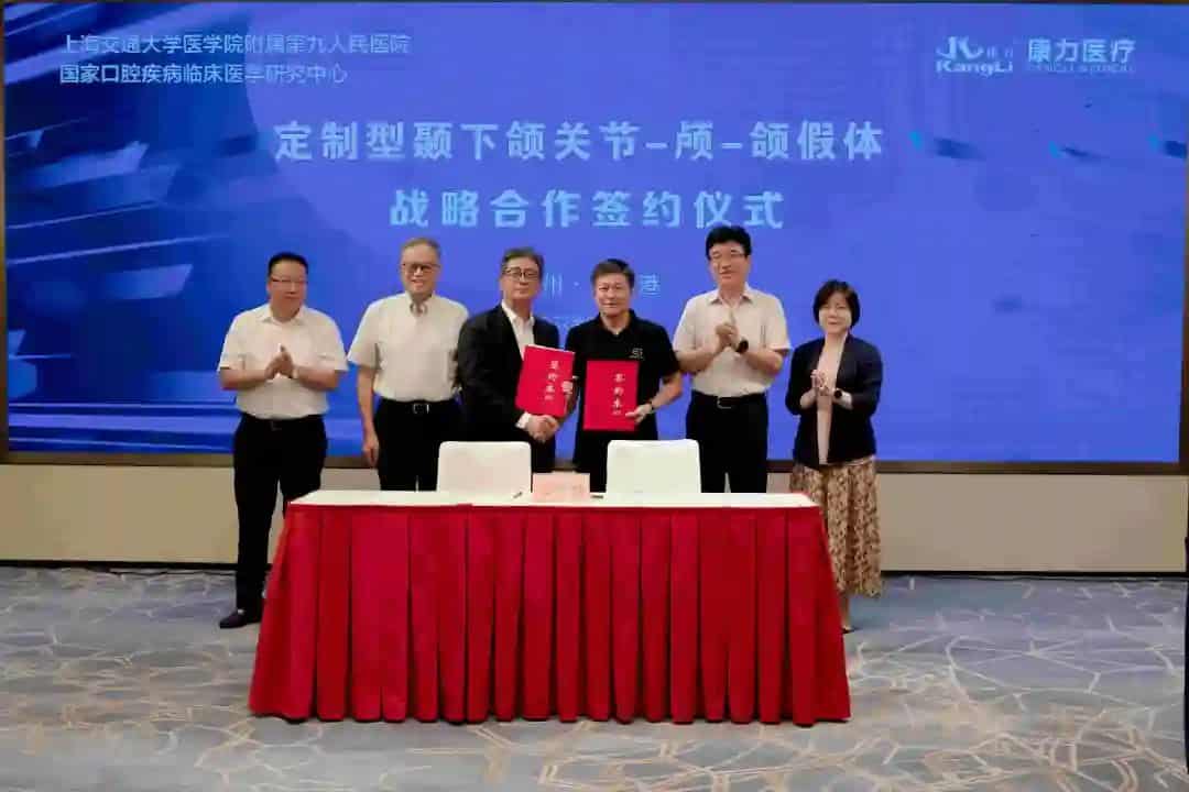 Six participants stand behind a table at a formal signing ceremony, with two red document folders, two handheld microphones and a large blue presentation screen behind them.