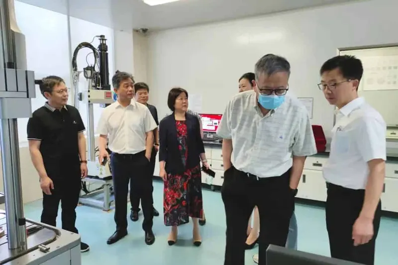 Seven participants stand around stainless-steel machinery, exposed tubing, workbenches, cabinets and computer equipment.
